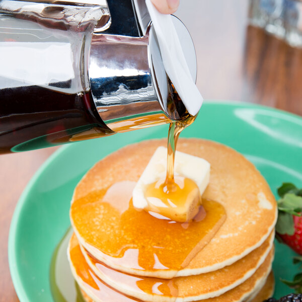 A glass syrup dispenser with a chrome-plated top pouring syrup onto a stack of pancakes with butter.