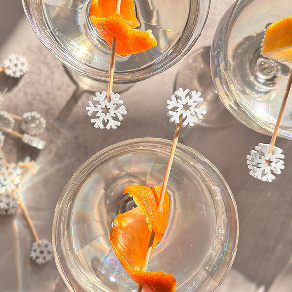 A set of clear drinks garnished with orange peels and decorated with bamboo cocktail picks featuring white snowflake designs.
