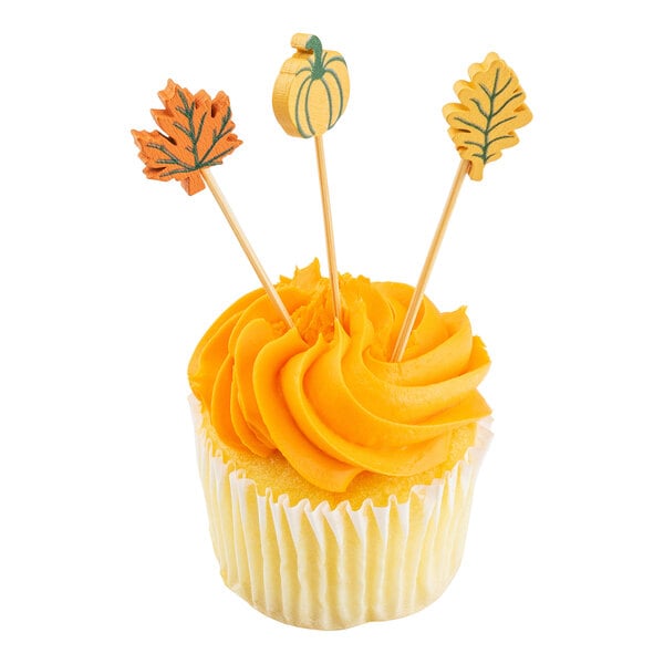 A cupcake decorated with orange frosting and three bamboo cocktail picks featuring fall leaves and a pumpkin design.