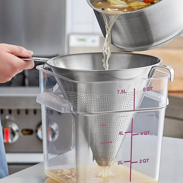 A stainless steel conical strainer being used to strain liquid from a pot into a clear plastic container.