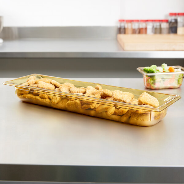 A Vollrath amber plastic food pan filled with chicken nuggets on a counter with a bowl of vegetables.