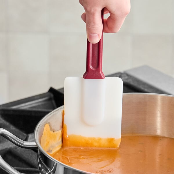 A hand is using a white silicone spatula with a red handle to stir orange sauce in a stainless steel pot on a stove.