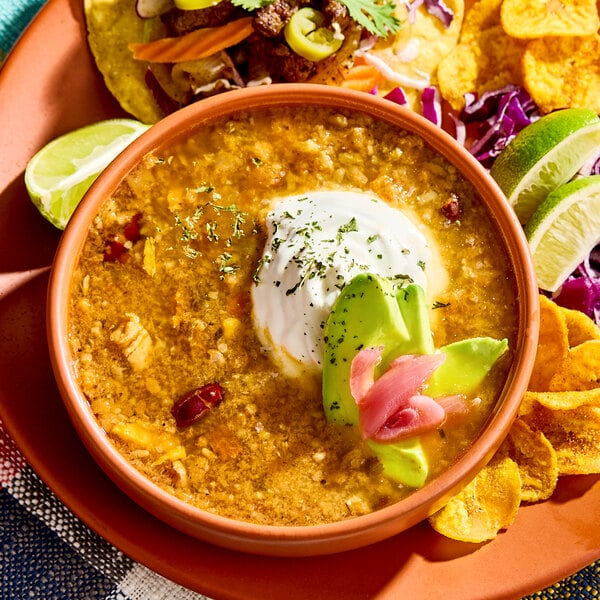 A bowl of Caribbean jerk chicken soup garnished with sour cream, avocado slices, and pickled onions, served with lime wedges and plantain chips.