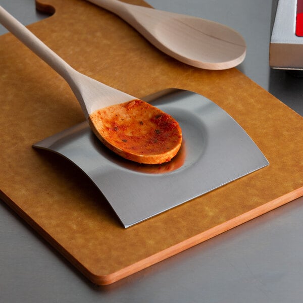 A brushed stainless steel square spoon rest holding a wooden spoon with sauce on a cutting board.