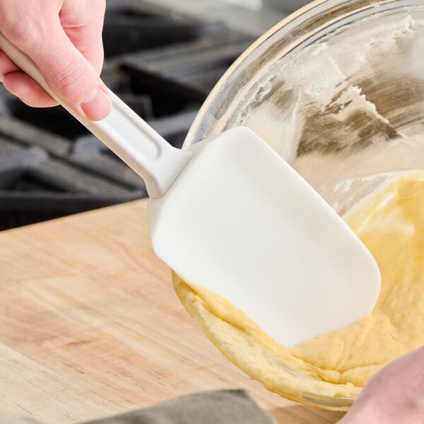 A person is using a white rubber spatula to scrape dough from a glass mixing bowl.