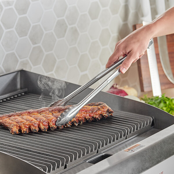 A pair of 16-inch extra heavy-duty stainless steel utility tongs being used to handle ribs on a grill.