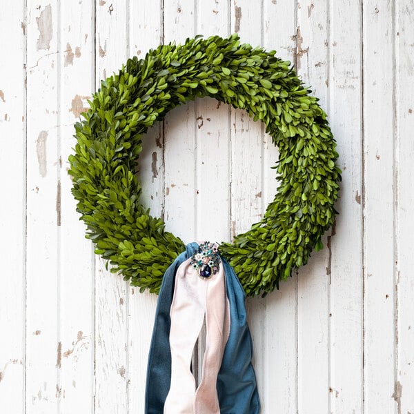 A round preserved boxwood wreath with green leaves, decorated with blue and pink ribbons and a jeweled accent, hanging on a rustic white wooden background.
