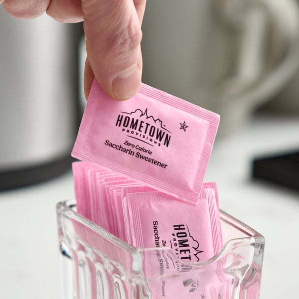 A pink packet of Hometown Provisions zero calorie saccharin sweetener being picked up from a glass container filled with similar packets.