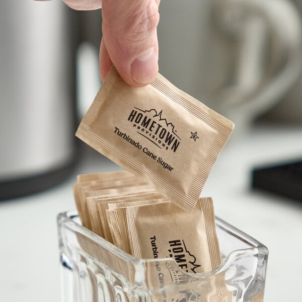 A small brown packet of Hometown Provisions turbinado cane sugar being picked up from a glass container.