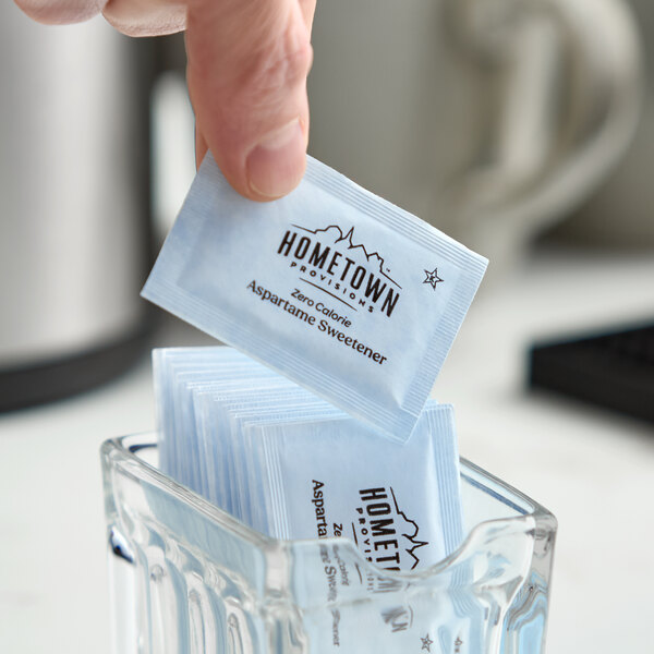 A blue packet of Hometown Provisions aspartame sugar substitute being picked up from a glass container.