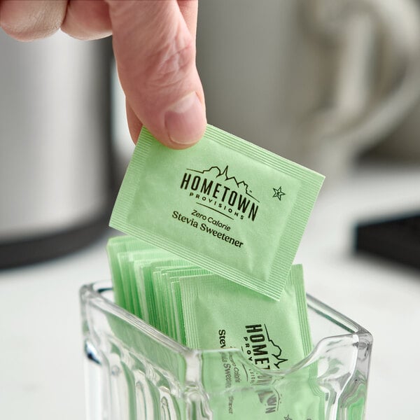 A green packet of Hometown Provisions zero calorie stevia sweetener being picked up from a glass container filled with similar packets.