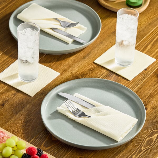 A cream-colored, linen-feel paper napkin folded on a plate with cutlery, set on a wooden dining table.