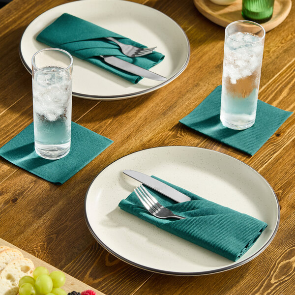 A set of hunter green airlaid linen-feel dinner napkins folded on plates and used as coasters on a wooden dining table.