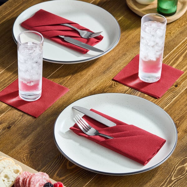 A set of burgundy airlaid linen-feel beverage napkins placed under glasses and used to hold cutlery on a dining table.