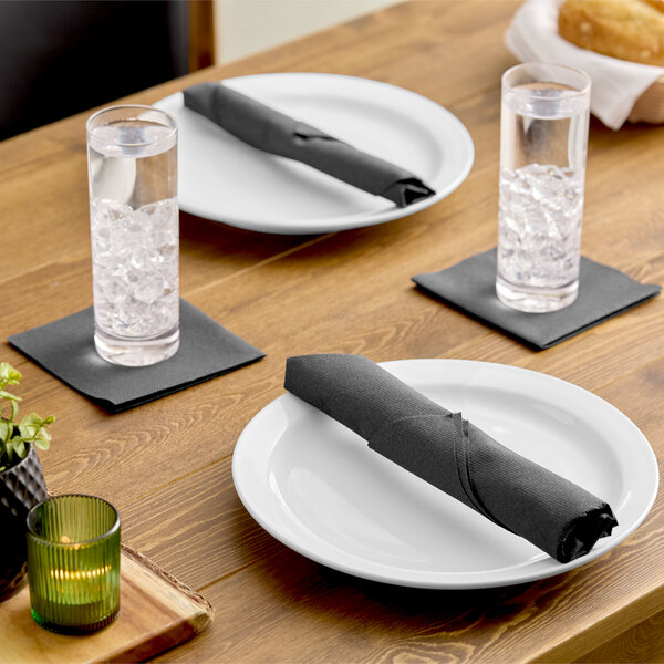 A set of white plates with black linen-feel dinner napkins, accompanied by glasses of ice water on black napkins, arranged on a wooden table.