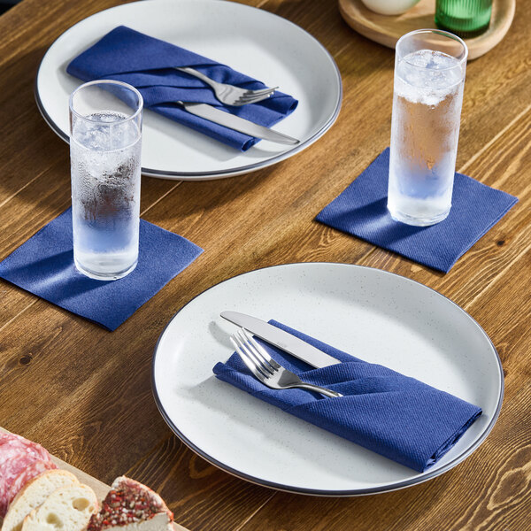 A set of navy blue linen-feel beverage napkins placed under glasses and holding cutlery on a wooden dining table.
