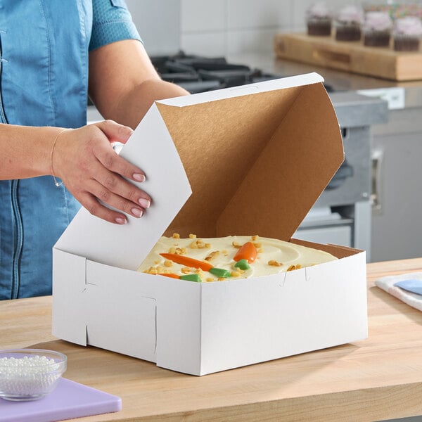 A white, customizable bakery box containing a cake, being closed by a person in a kitchen setting.
