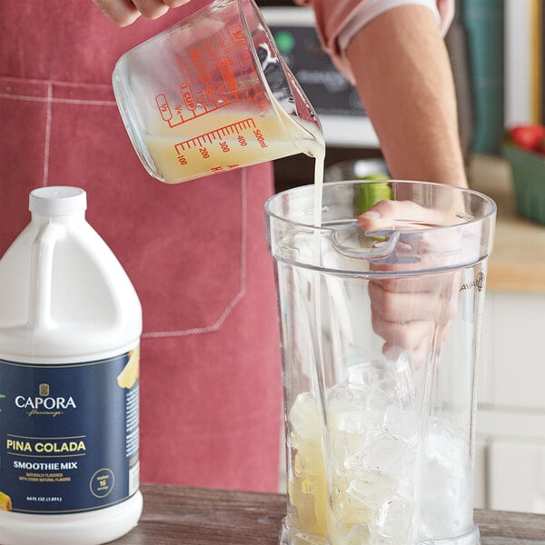A bottle of Capora Pina Colada Fruit Smoothie Mix next to a blender being filled with the mix and ice.