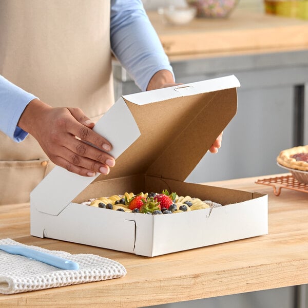 A white, square bakery box being opened to reveal a fruit-topped pie inside.