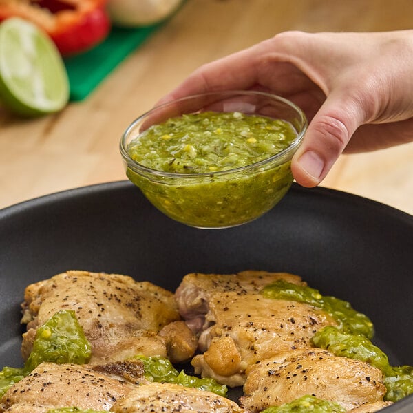 A small glass bowl of green sofrito sauce being held above a pan of cooked chicken.