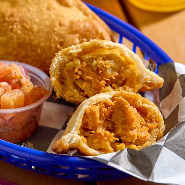 A buffalo chicken empanada with a flour shell, cut open to show the filling, served in a basket with a side of salsa.