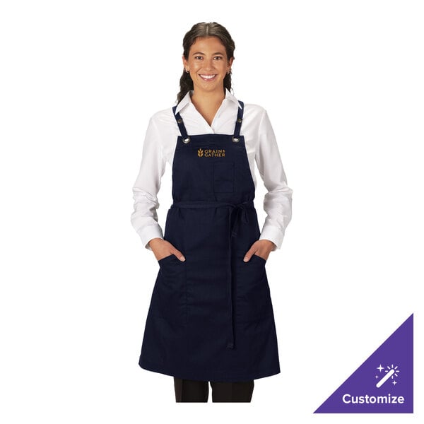 A navy blue customizable poly/cotton cross-back bib apron with three pockets, measuring 34 by 28 inches.