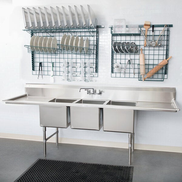 A large stainless steel three-compartment commercial sink with two drainboards, stainless steel legs, and cross bracing, installed in a kitchen setting.