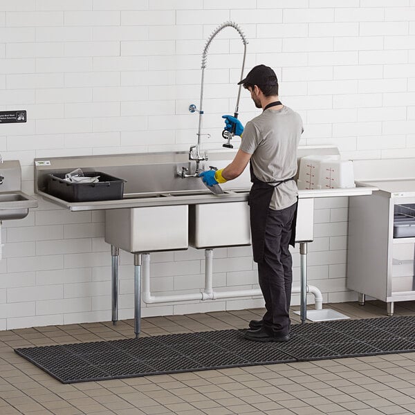 A stainless steel three compartment commercial sink with two 24-inch drainboards and galvanized steel legs, shown in use in a commercial kitchen setting.