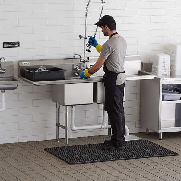 A stainless steel two-compartment commercial sink with two drainboards, stainless steel legs, and cross bracing, shown in use in a commercial kitchen setting.