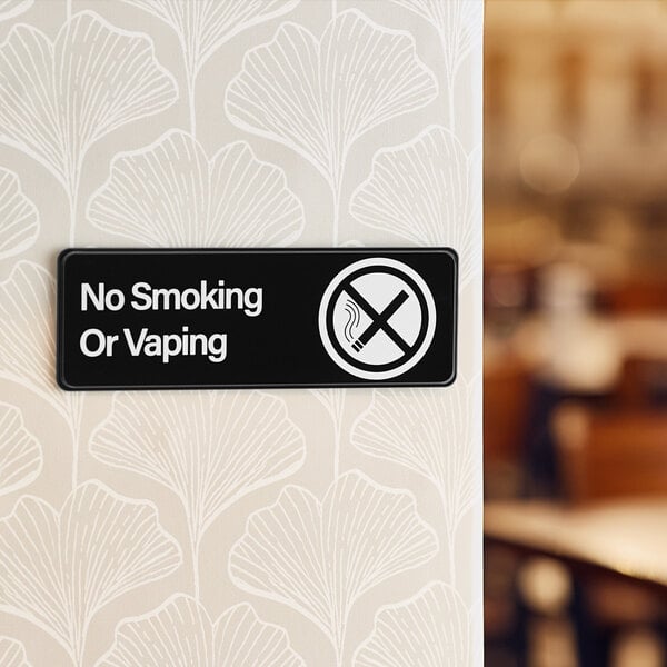 A rectangular black and white sign that reads 'No Smoking Or Vaping' with a crossed-out cigarette symbol.