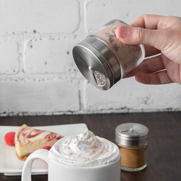 A small glass spice shaker with a stainless steel adjustable lid being used to sprinkle powder onto a mug of whipped cream.