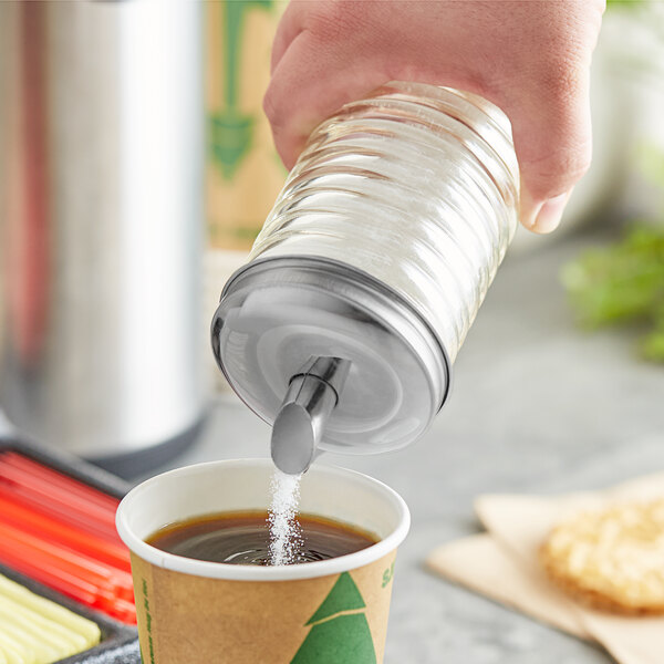 A glass sugar pourer with a stainless steel spout being used to pour sugar into a cup of coffee.