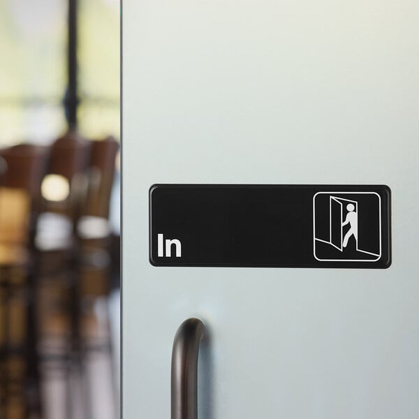 A black and white plastic sign on a door indicating entry with the word 'In' and a pictogram of a person entering.