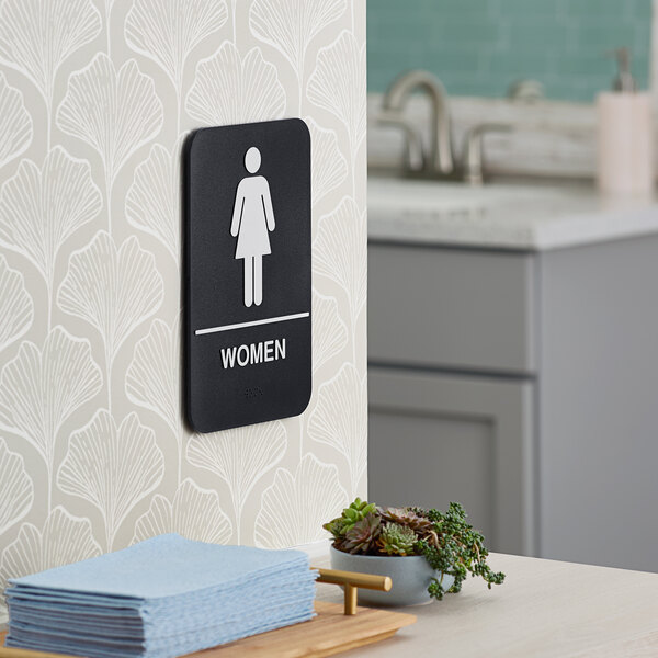 A black and white plastic women's restroom sign with braille mounted on a wall.