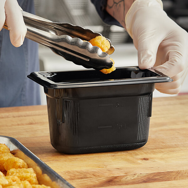 A person using tongs to serve food from a Vollrath black plastic food pan on a wood surface.