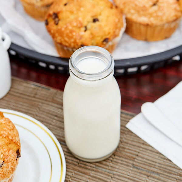 A small clear glass milk bottle filled with milk, placed on a table with muffins and a plate nearby.