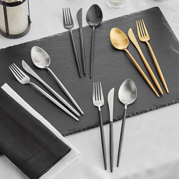 A 5-piece set of Acopa Odin flatware in black, silver, and gold finishes, displayed on a slate serving board.