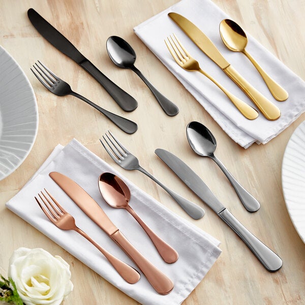 A set of five-piece flatware in various finishes, including black, gold, silver, and copper, displayed on white napkins on a wooden table.