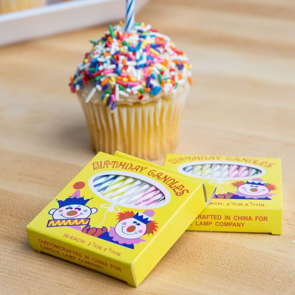 A box of multi-colored birthday candles with clown illustrations on the packaging, shown next to a cupcake with sprinkles.