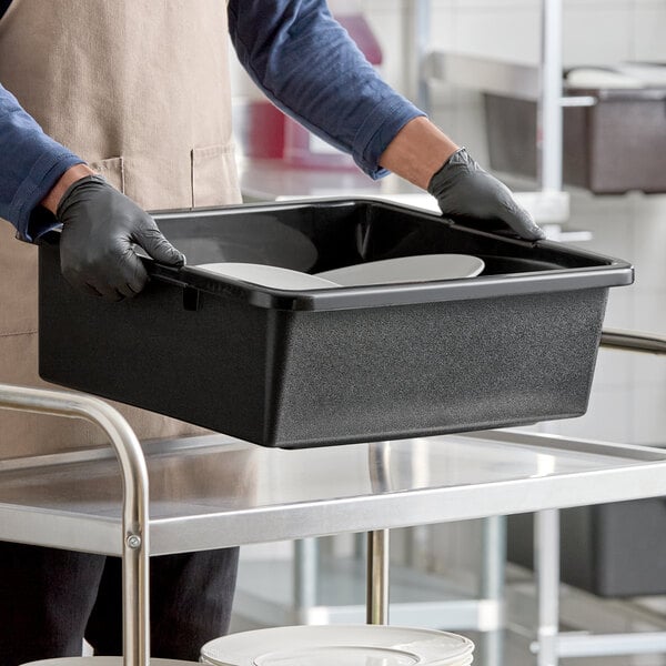 A black heavy-duty polyethylene bus tub or tote box being used to carry dishes in a commercial kitchen setting.