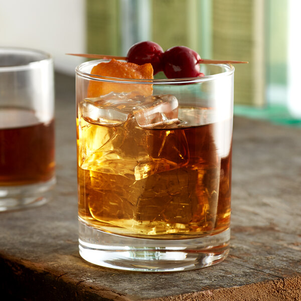 A glass of whiskey on the rocks garnished with cherries and an orange peel, with a wooden toothpick holding the garnish.