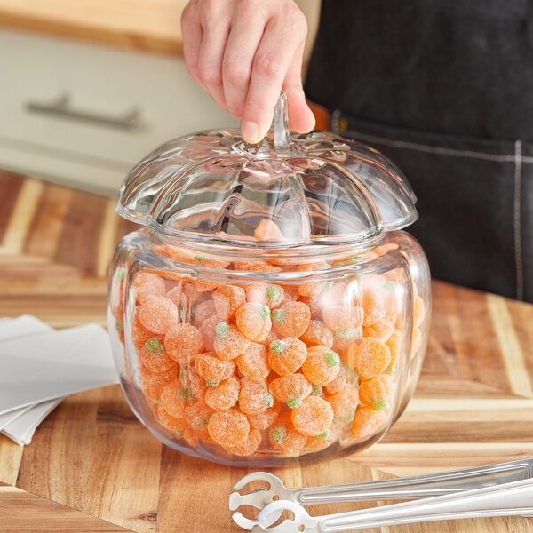 A clear glass pumpkin-shaped jar with a lid, filled with orange pumpkin-shaped candies.