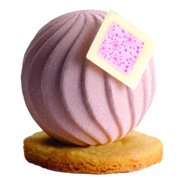 A pink spherical chocolate dessert with a textured surface, decorated with a white and pink square, sitting on a round cookie base.