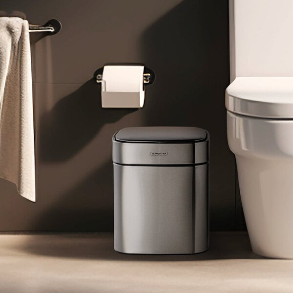 A stainless steel square wastebasket with an automatic lid, placed next to a toilet in a bathroom.