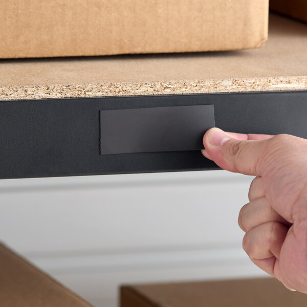 A hand placing a rectangular pre-perforated magnetic label holder on the edge of a steel shelf.