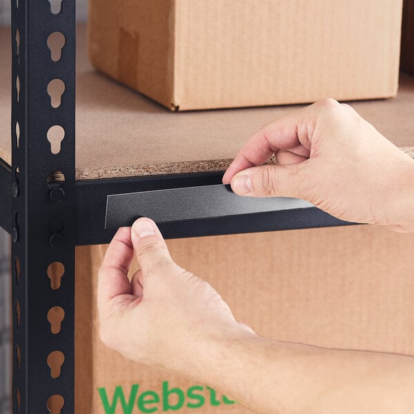 A black 1-inch by 36-inch slip strip label holder being applied to a metal shelf.