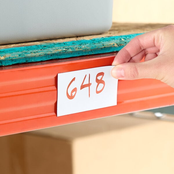 A white magnetic write-on label with the number 648 written on it, being placed on an orange metal shelf.