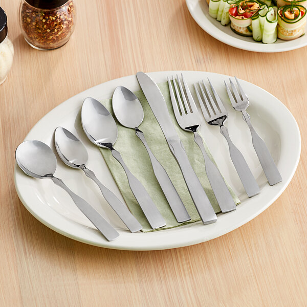 A set of stainless steel flatware including spoons, forks, and a knife displayed on a white oval plate.