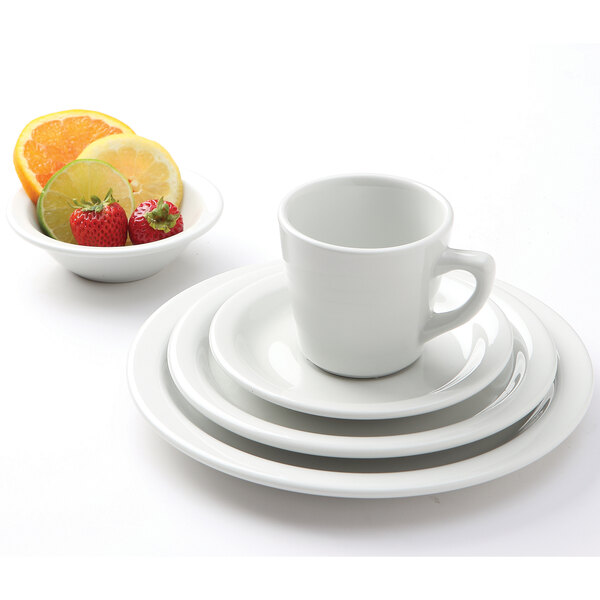 A white Tuxton tall cup on a saucer with a bowl of fruit including strawberries and lime slices.