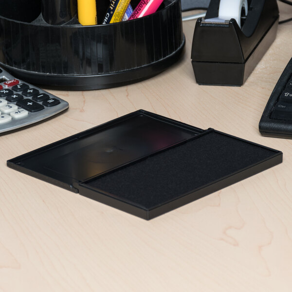 A black pre-inked felt stamp pad in a rectangular plastic case, shown open on an office desk.